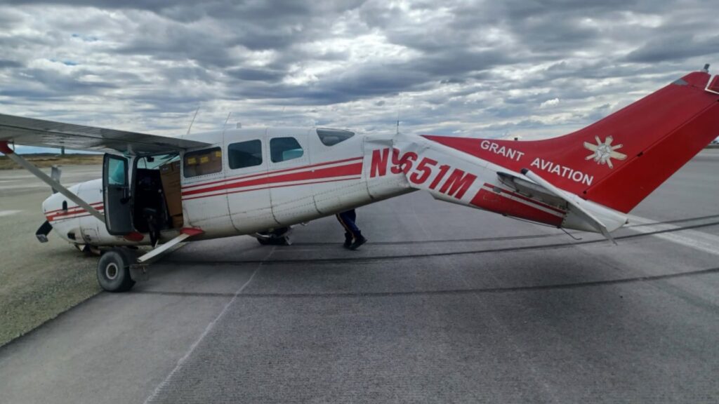 A damaged Cessna 207A aircraft with the registration number N9651M is shown on a tarmac under a cloudy sky. The fuselage is visibly bent and creased near the empennage, indicating substantial damage from a hard landing. The aircraft bears the "GRANT AVIATION" logo on the red tail, and the cargo door is open with boxes visible inside. A person is partially visible beneath the aircraft.