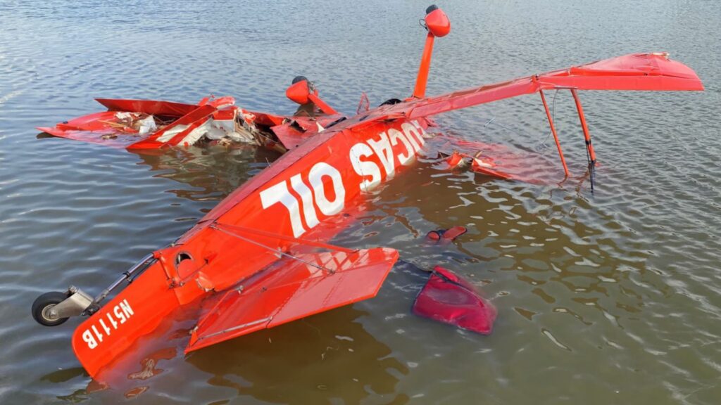 Wreckage of a red Aviat S-1-11B aircraft upside down and partially submerged in a river, with visible damage to the fuselage and wings. The tail number N5111B and “LUCAS OIL” branding are visible.