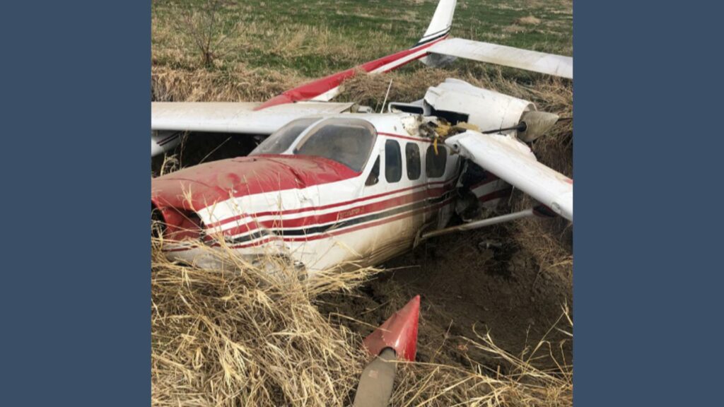 Front and side view of the wrecked Cessna P337H N62PC in a field, with engine damage and detached propeller hub lying in dry grass.