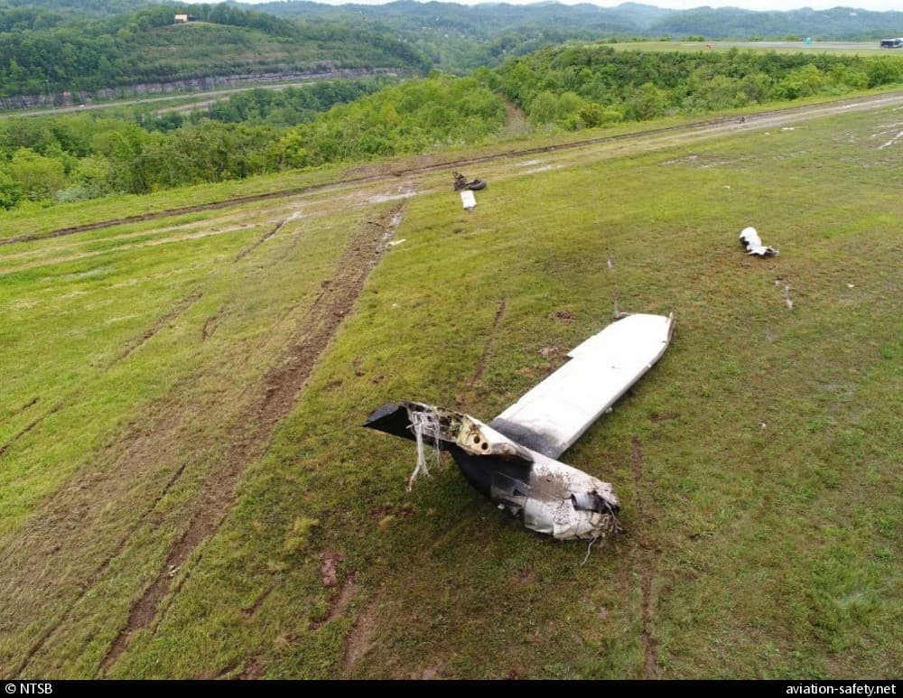 Scattered aircraft wreckage on a grassy slope near an airport runway, with a detached wing in the foreground and debris trail leading downhill into a wooded valley.