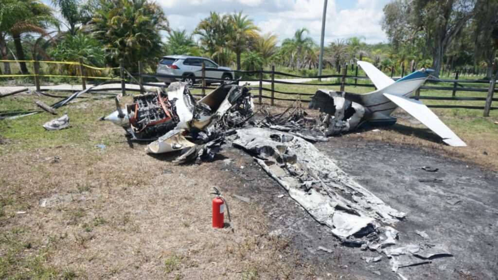 Burned wreckage of a Commander 114TC aircraft, showing a charred engine, destroyed fuselage, and wing remains, with a fire extinguisher in the foreground and fencing in the background.