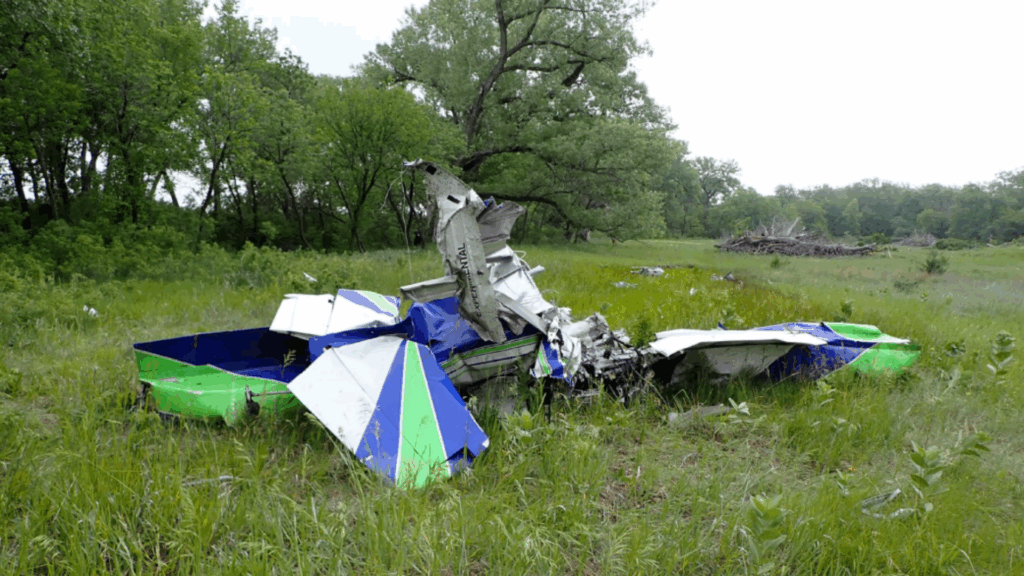 Wider view of the crash site showing the destroyed Van’s RV-7 aircraft wreckage lying in a grassy field surrounded by trees. The vertical stabilizer marked 'EXPERIMENTAL' is visible amid the wreckage.