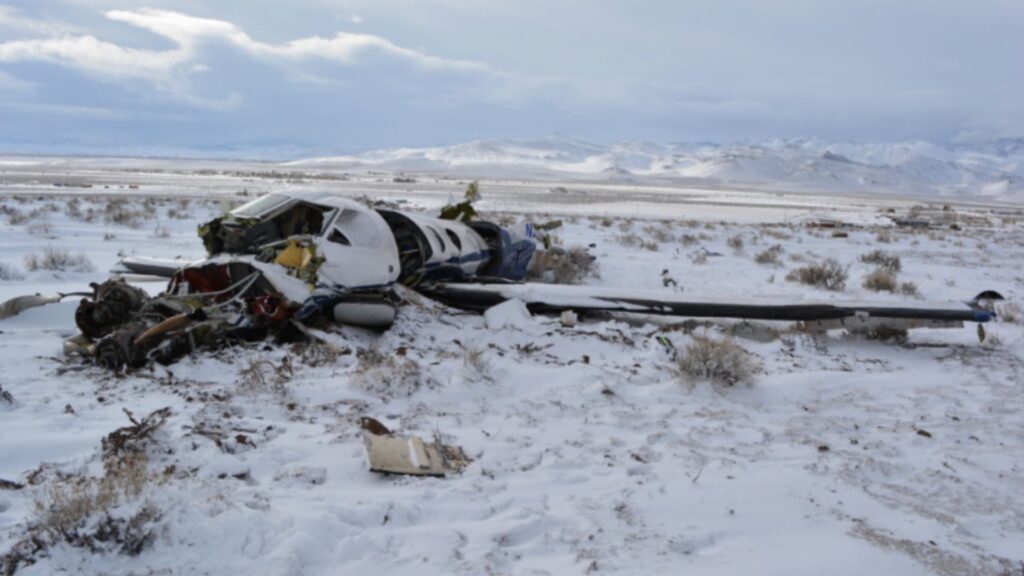 Wreckage of a Pilatus PC-12 aircraft (registration N273SM) resting in a snow-covered desert near Stagecoach, Nevada. The fuselage is heavily damaged and partially crushed, with one wing detached and scattered debris surrounding the crash site. Snow-covered mountains are visible in the background under a cloudy sky.