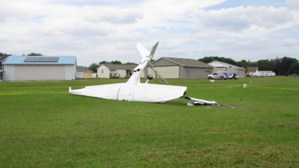 Crashed Merlin Lite motorglider N622PC lying inverted on a grassy airfield in Mulberry, Florida, with significant structural damage and emergency response vehicles in the background.