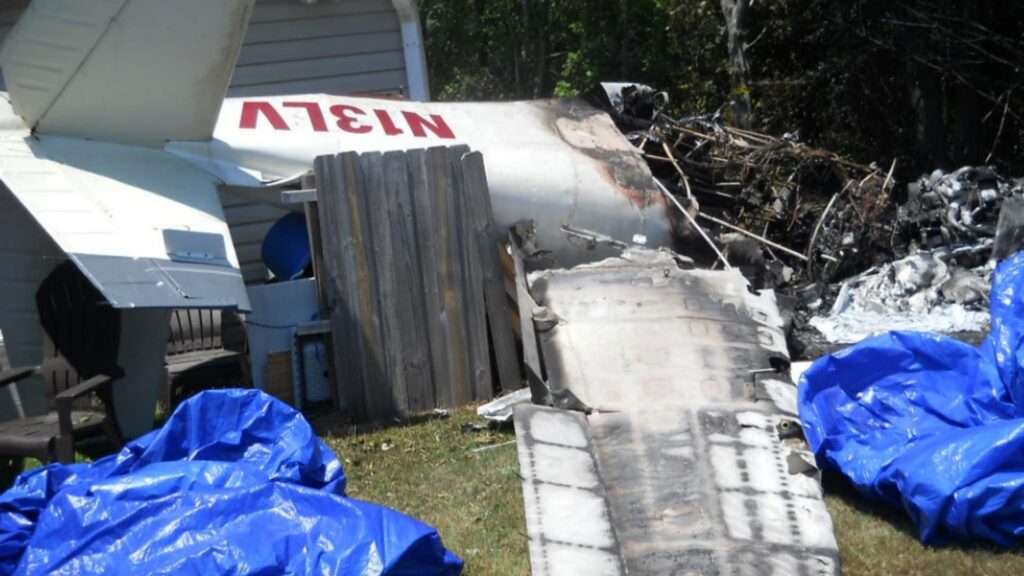 Burned aircraft wreckage, including a detached wing and the tail section marked with the registration number “N13LV,” lies against a house. Blue tarps and debris are scattered on the lawn.