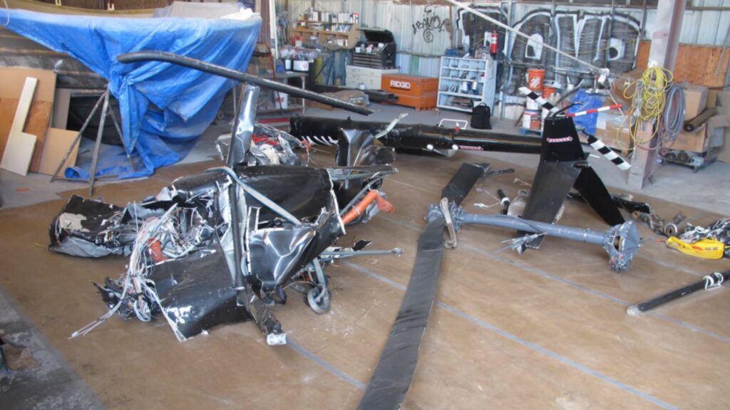 Wreckage of a Robinson R44 helicopter, including severely damaged fuselage, tail boom, and rotor components, laid out in a hangar for examination following a fatal in-flight breakup near Kauai, Hawaii.