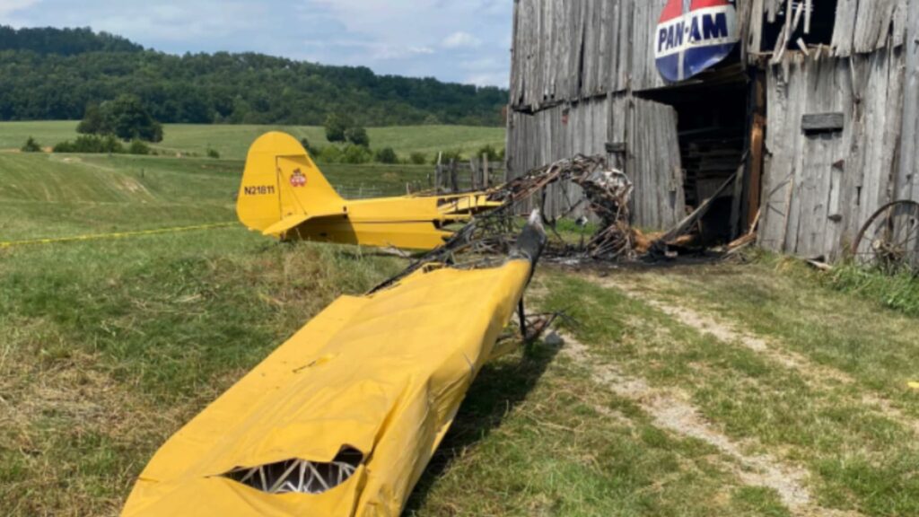Wreckage of a yellow Piper J3 Cub (N21811) lying in front of a weathered wooden barn with a large vintage Pan-Am sign. The aircraft’s wing is detached and damaged, and the fuselage is burned near the building's entrance.