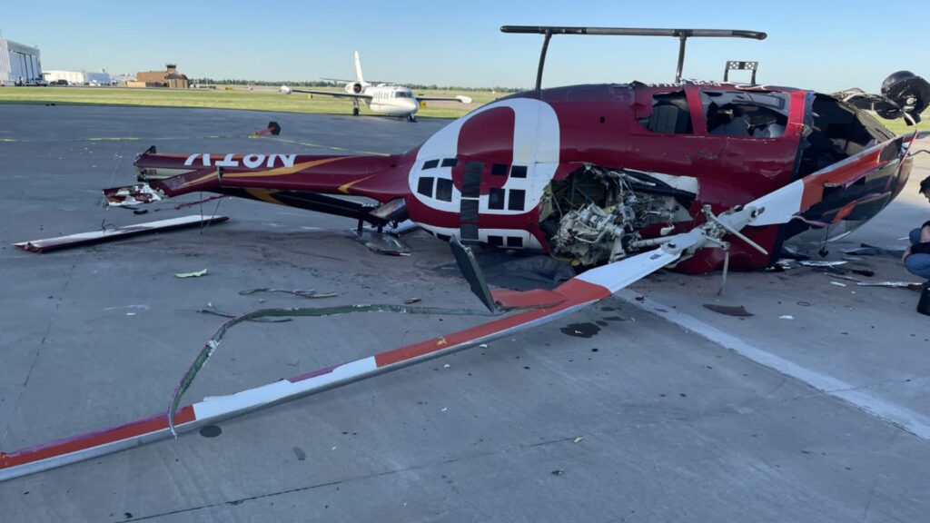Crashed red Bell 505 helicopter lying on its right side on a concrete airport ramp. The main rotor blades are severely damaged and bent, and the engine compartment is exposed with internal components visible. The tail boom is broken and twisted. A business jet and airport buildings are seen in the background under a clear blue sky.