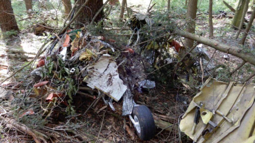 Close-up of fragmented wreckage of a Piper PA-30 in a wooded area, showing crumpled metal, wires, and landing gear amidst fallen branches.