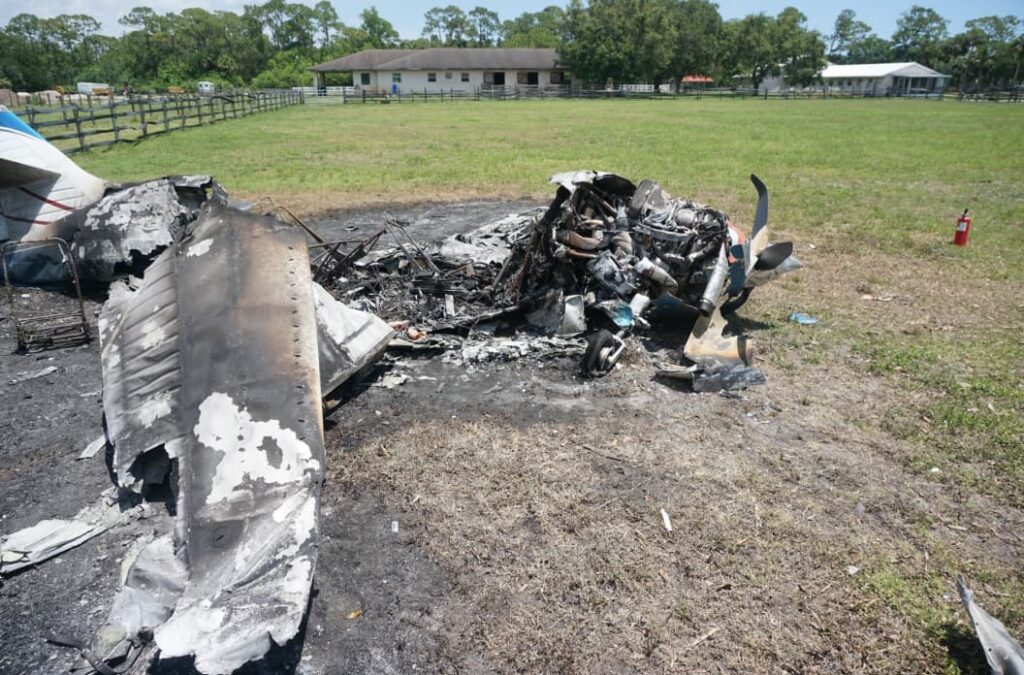 Close-up of the Commander 114TC burned engine and wing debris, resting on scorched earth, with rural buildings and fences visible in the distance.