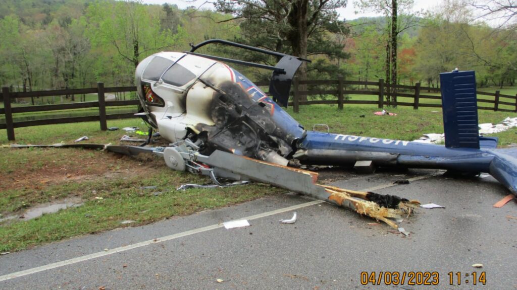 Wreckage of an Airbus EC130 T2 helicopter lying on its left side near a rural road, showing severe fuselage and tail damage, burn marks, and broken rotor blades, with a wooden fence and trees in the background.
