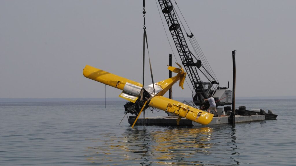 A yellow Avid Magnum amphibious airplane being hoisted from Lake Michigan by a crane mounted on a barge. The aircraft shows significant damage, with bent wings and a broken tail section. The calm water reflects the wreckage and the crane.