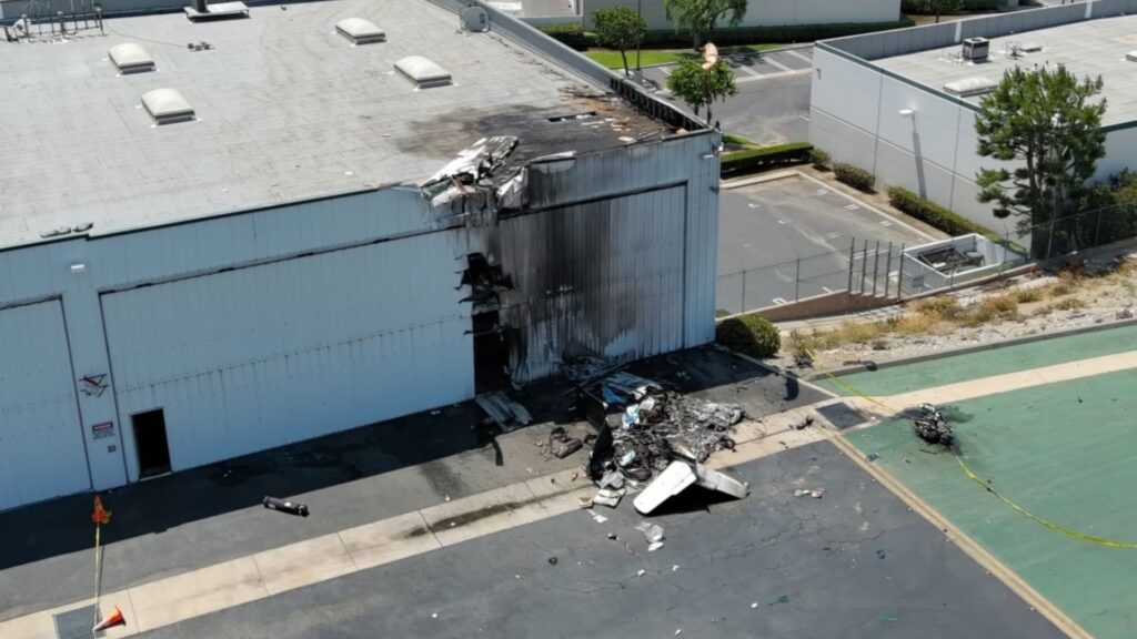 Aerial view of a large white hangar with a gaping hole and black fire damage where an aircraft struck the wall and roof. Burned wreckage lies on the ground near the building’s doorway.