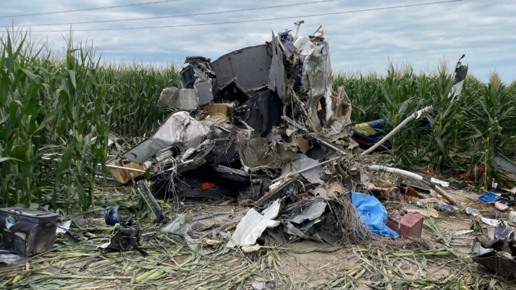 Wreckage of a UH-1H helicopter crash in a cornfield, showing severely mangled and fragmented fuselage debris surrounded by broken corn stalks. The crash site is located under cloudy skies with visible power lines overhead.
