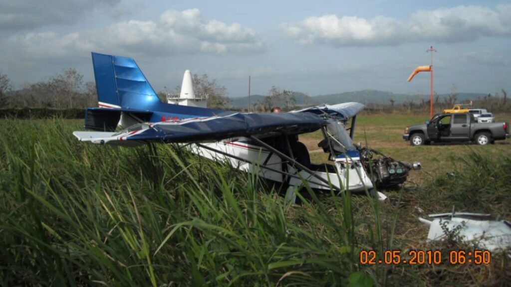 Side view of the wrecked RANS S-6S in grass near the runway, bent left wing, crushed nose, and a pickup truck in the background.