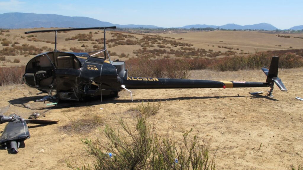 Wreckage of Robinson R22 helicopter N7527Y resting on its left side in a dry, brush-covered area near Murrieta, California. The tail boom remains mostly intact while the main rotor is detached and lying on the ground nearby.