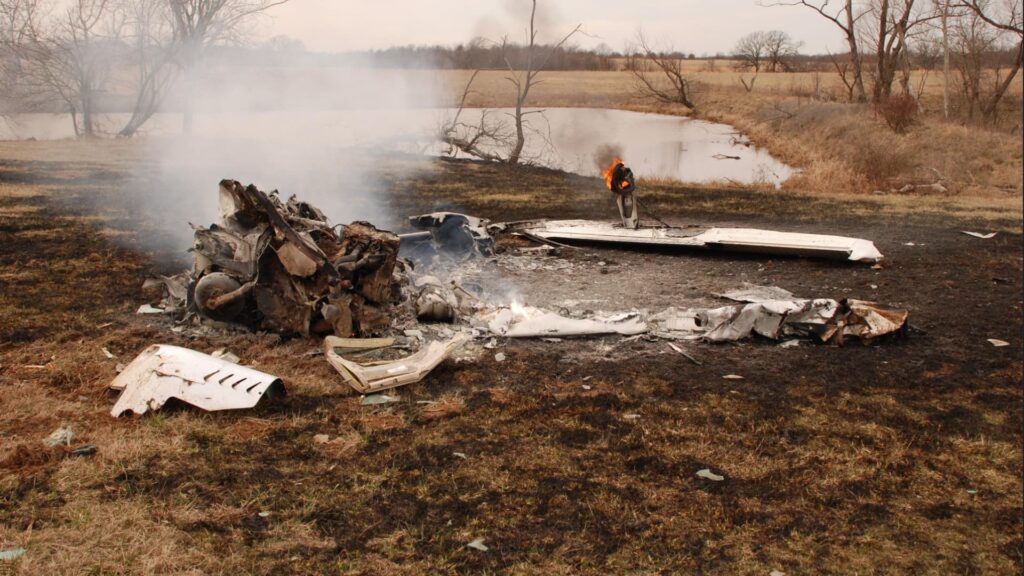 Burned wreckage of a small aircraft scattered in a grassy field near a pond, with smoke rising from the debris and a small fire still burning on part of the structure.