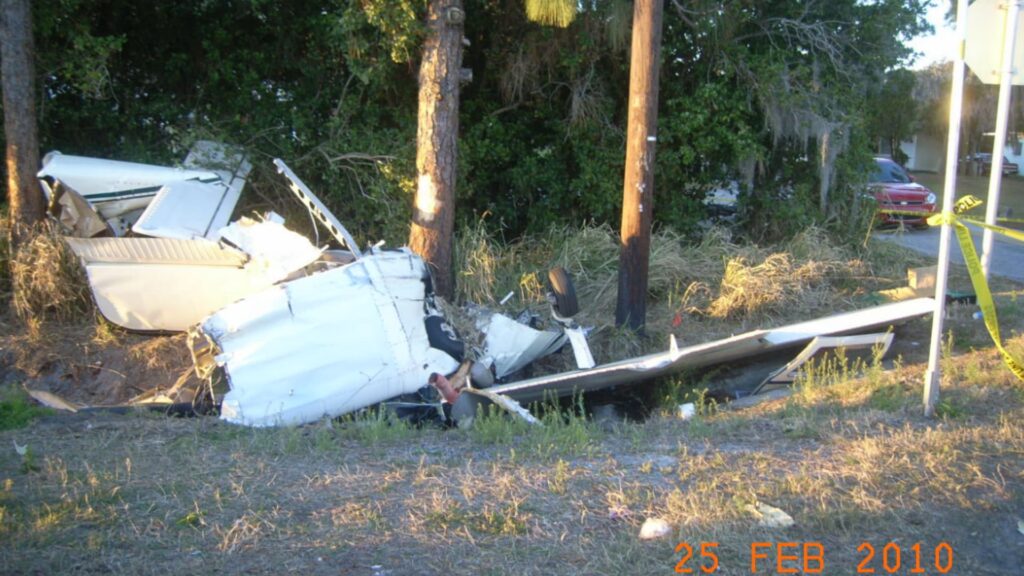 Wreckage of a Beech C23 airplane lying in a ditch beside US-92 in Winter Haven, Florida. The fuselage and wing are broken apart near a tree and utility pole, with debris scattered in the grass. A street sign for Pickney Drive and US-92 is visible.