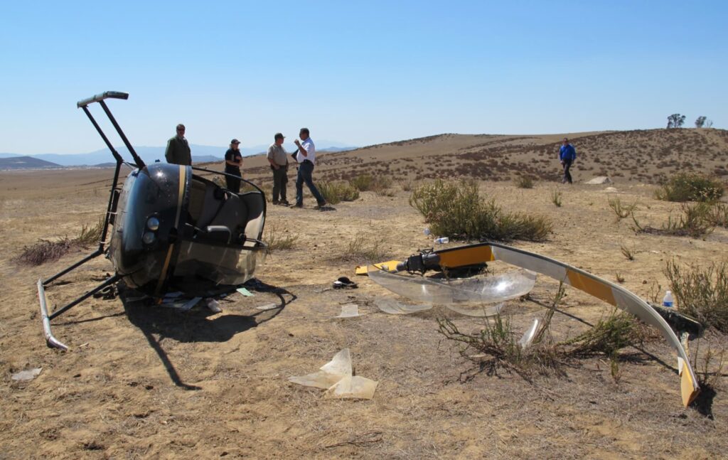 Frontal view of overturned Robinson R22 helicopter N7527Y at the crash site. The cockpit is heavily damaged and scattered debris, including a broken main rotor blade and plexiglass windshield, lies across the arid terrain.