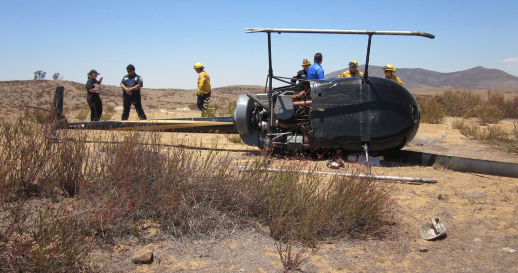Side view of Robinson R22 N7527Y wreckage with the tail boom stretched out and engine components exposed. Firefighters and investigators are present at the site, surrounded by dry brush and hilly terrain.