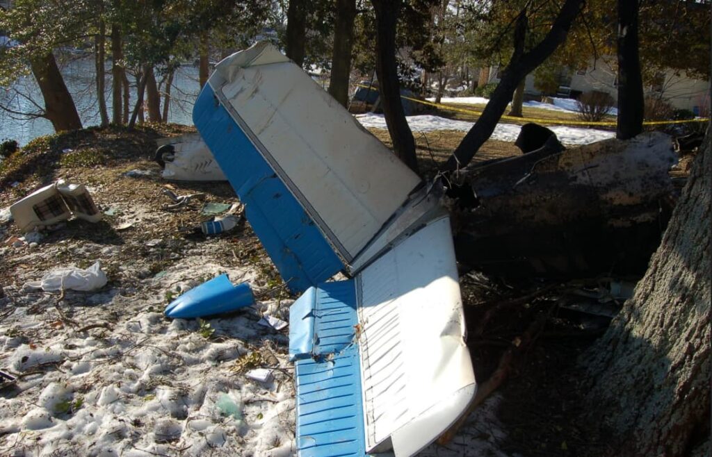 Detached tail section of the airplane with blue-and-white paint, broken off and leaning against trees.
