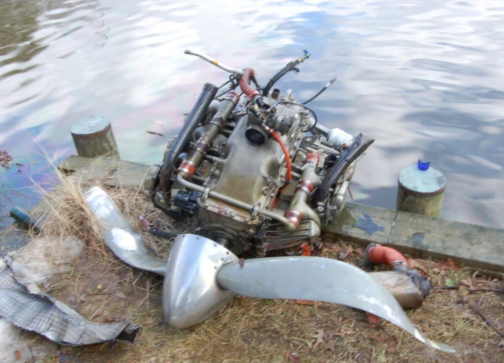 Aircraft engine with a bent two-blade propeller resting on the edge of a creek.