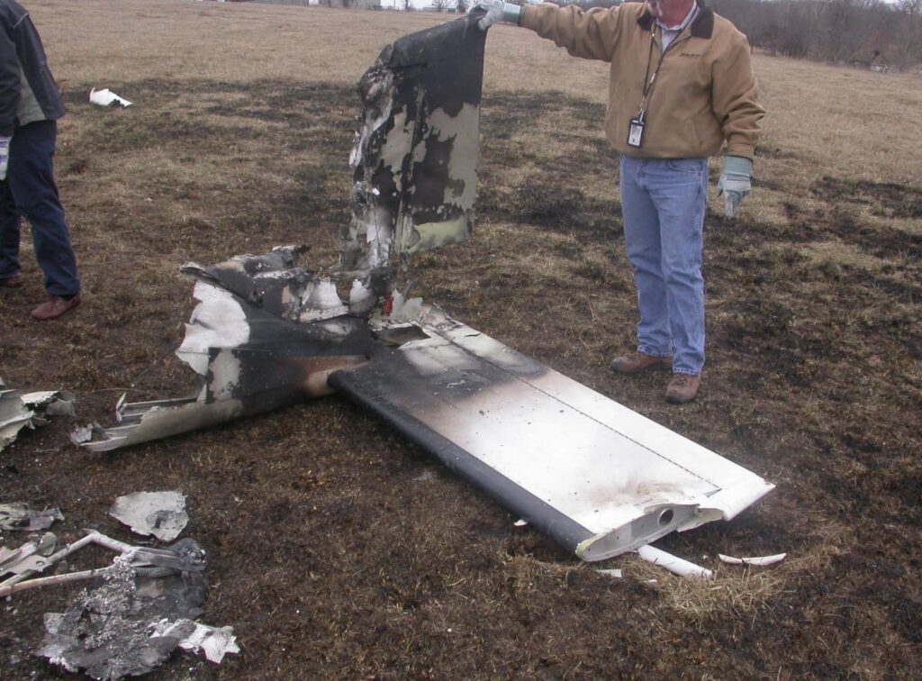An investigator in jeans and gloves holds up a charred section of the aircraft’s tail assembly, showing significant fire damage to the horizontal and vertical stabilizers.