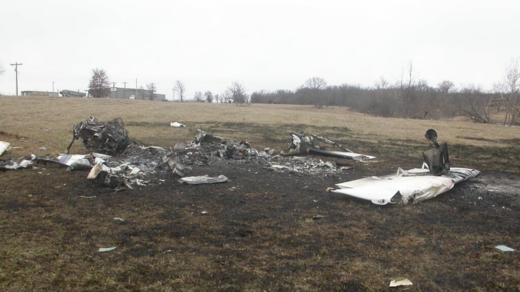 Wide view of the crash site showing multiple burned and twisted sections of the airplane wreckage spread across a blackened patch of grass.