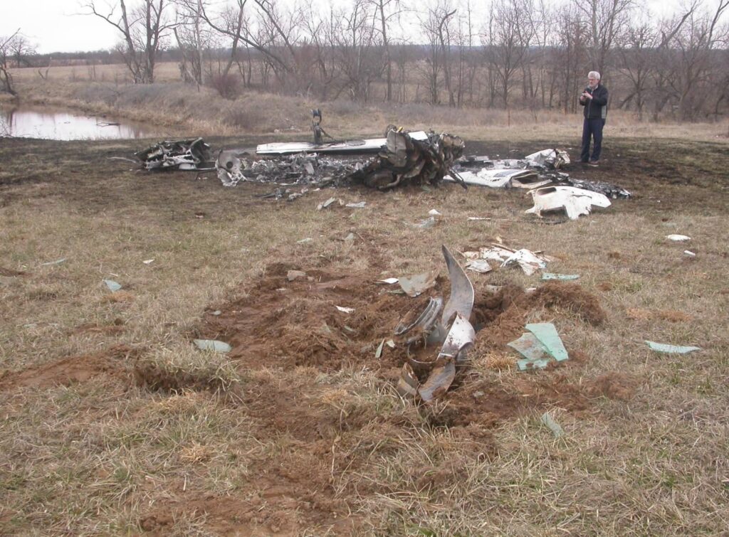 Ground-level view of the crash site with debris scattered across scorched grass; a broken propeller blade is visible in the foreground, with the burned fuselage in the background near a pond.