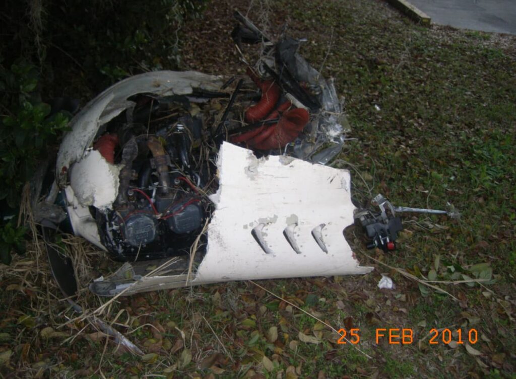 Severely damaged aircraft engine from the Beech C23 resting on the ground. The engine cowling is torn away, exposing cylinders, wiring, and red hoses, with broken sheet metal around it.
