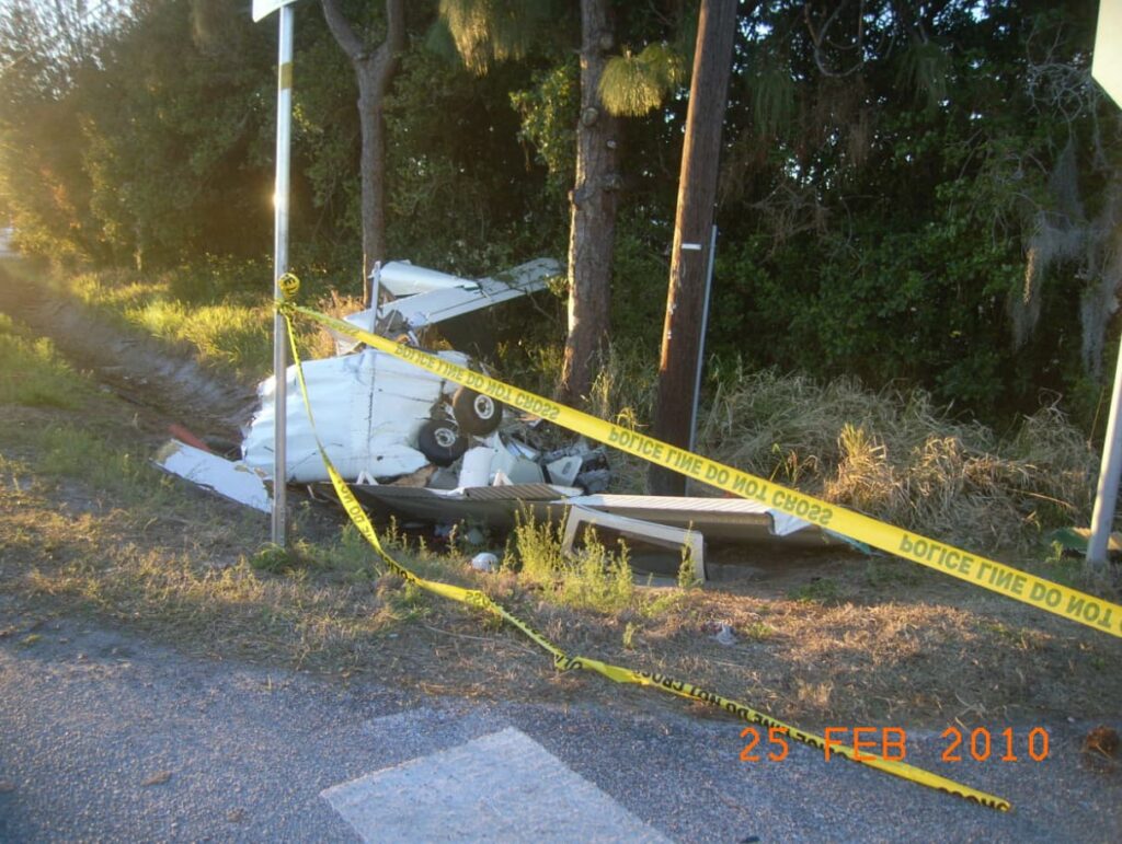 Crashed Beech C23 wreckage in a grassy ditch next to a roadway. Yellow police tape surrounds the debris, which includes part of the fuselage and wing structure, resting against a tree and utility pole.