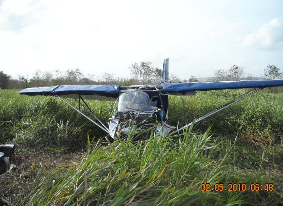 A small blue-and-white RANS S-6S aircraft wrecked in tall grass, nose crushed, wings bent, and resting inverted on uneven terrain.
