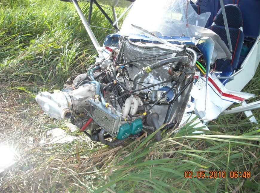 Close-up of the damaged Rotax engine pulled forward from the crumpled fuselage, wires and hoses exposed.