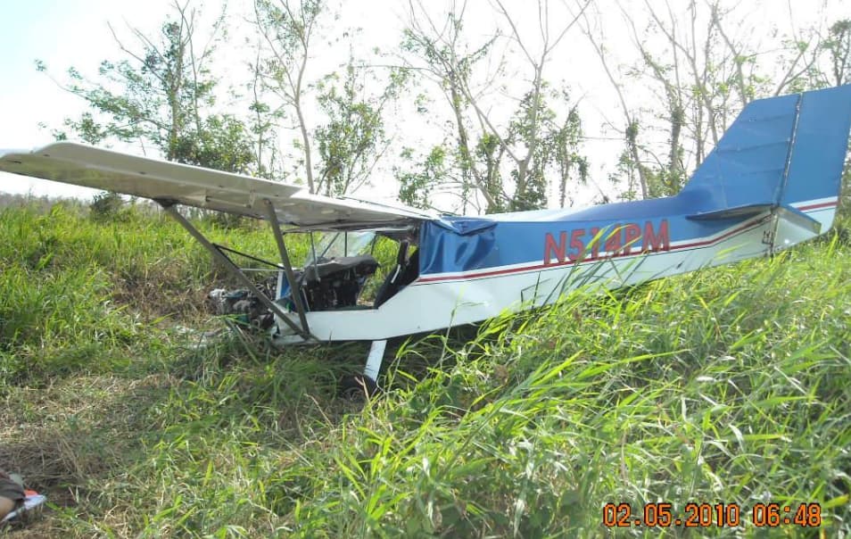 Rear-left view of the crashed aircraft showing tail number N514PM, bent fuselage, collapsed landing gear, and destroyed engine section.