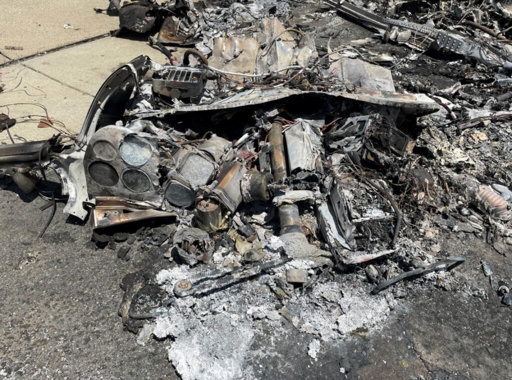 Close-up of heavily burned and melted cockpit instruments, wiring, and structural components from the crashed aircraft, lying in a pile of charred debris on the pavement.