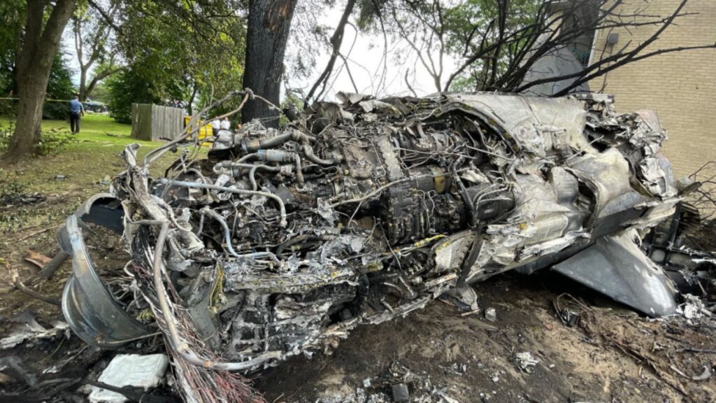 Wreckage of a burned and twisted aircraft engine lying against a tree and building wall, with charred debris, exposed wires, and crushed metal components visible.