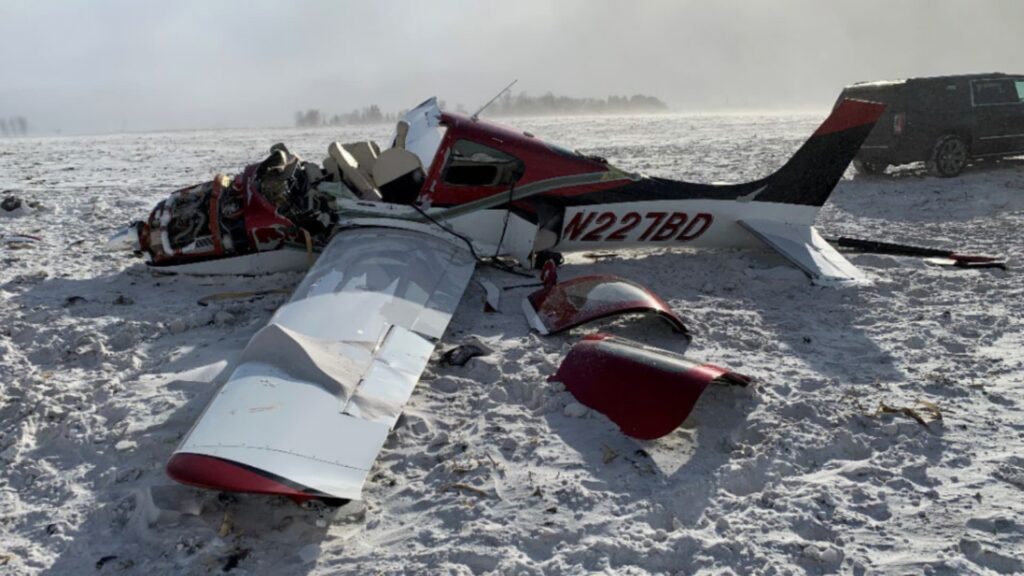 A heavily damaged red and white Cirrus SR22, registration N227BD, lies in a snowy field after a crash. The forward fuselage is crushed with the engine exposed, and both wings are broken with debris scattered around. A black SUV is parked nearby under a hazy winter sky.