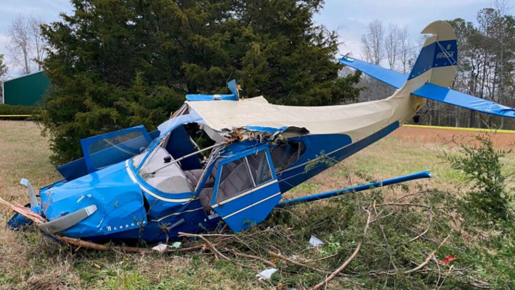 Wrecked blue-and-cream tailwheel airplane resting in a grassy field with broken wings and nose damage after colliding with trees; the registration number N6665K is visible on the tail.