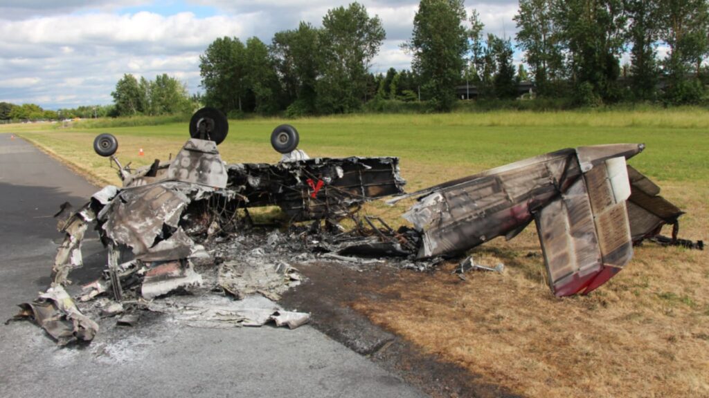 Burned wreckage of an inverted Beechcraft Bonanza aircraft resting on a runway and grass shoulder. The airframe is mostly destroyed by fire, with only the landing gear, wing fragments, and charred fuselage remaining. Trees and cloudy skies are visible in the background.