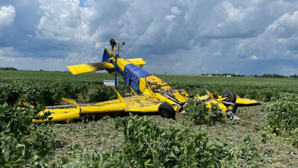 Closer ground-level view of the wrecked yellow agricultural airplane resting inverted in a soybean field. The fuselage is crushed, wings twisted, and a blue tarp partially covers the wreckage.