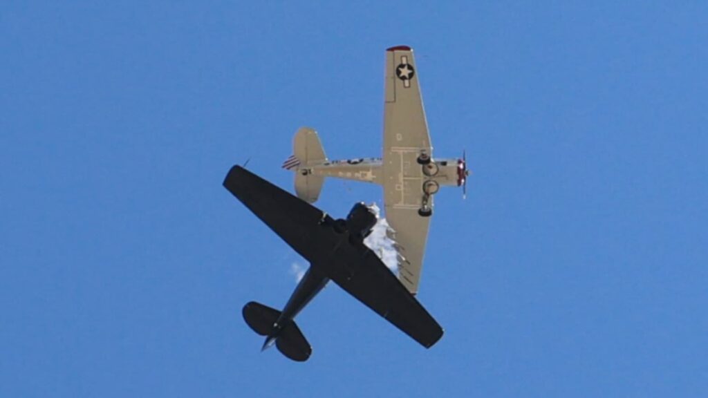 Two vintage North American T-6 aircraft collide midair against a clear blue sky, one painted black and the other in tan with U.S. Air Force markings, as debris and smoke appear from the impact.