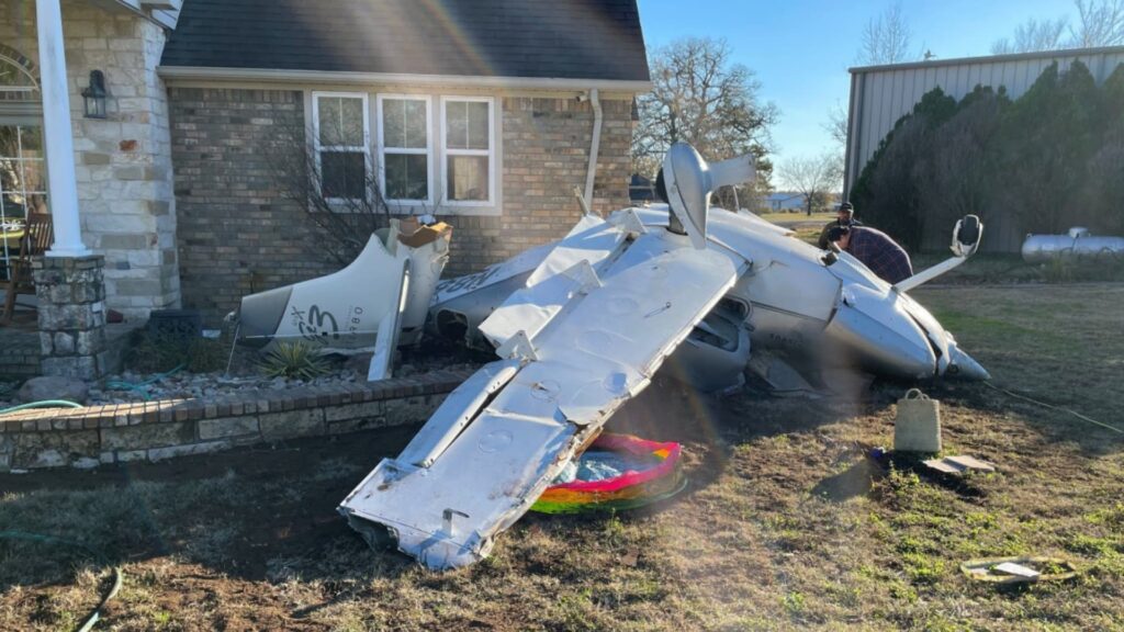 Crashed small aircraft lying upside down against the side of a brick house, with wings and fuselage heavily damaged; two people are standing nearby inspecting the wreckage.