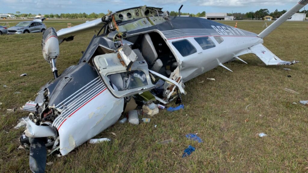 Close-up of the wrecked Piper PA-32-260 lying inverted in grass. The nose and cockpit area are crushed, with the windshield destroyed and debris scattered around. The fuselage is buckled but the tail remains attached, with hangars and cars visible in the background.