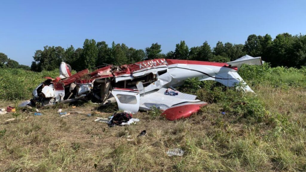 Wreckage of a red and white Piper PA-32 aircraft resting in a grassy field, heavily damaged with the fuselage crushed, wings broken, and debris scattered around.
