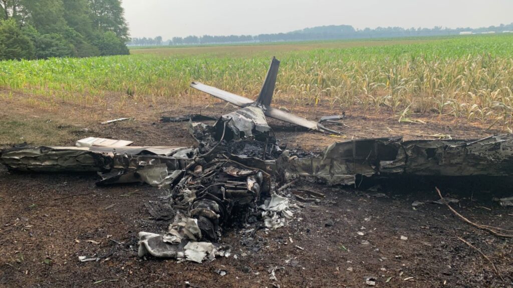 Burned wreckage of a small aircraft lying in a field beside a corn crop. The fuselage and wings are heavily charred and twisted, with the tail section partially intact and pointing upward. The scorched ground shows signs of fire damage surrounding the crash site.