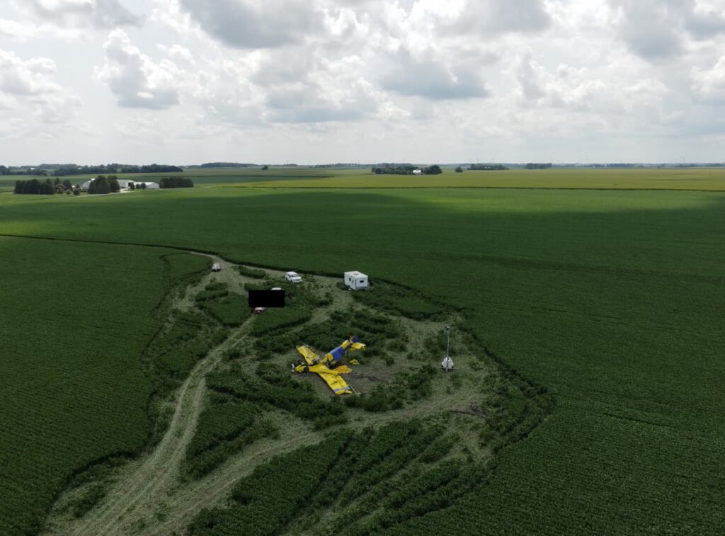 Aerial view of a yellow agricultural airplane that crashed in a green soybean field near Oxford, Indiana. The aircraft lies inverted and broken within a cleared section of crops, surrounded by several vehicles and equipment trailers.