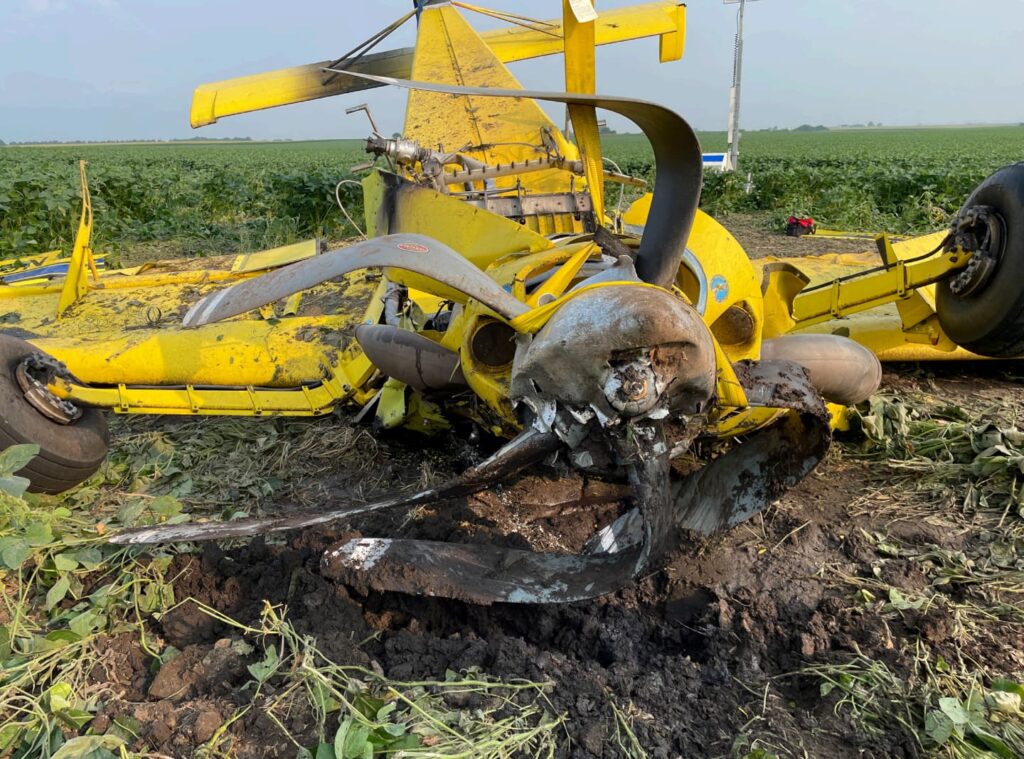 Detailed view of the Air Tractor’s damaged propeller and nose section. The bent and twisted propeller blades are embedded in the dirt, showing signs of power at impact. The wreckage of the fuselage and landing gear is visible behind it.