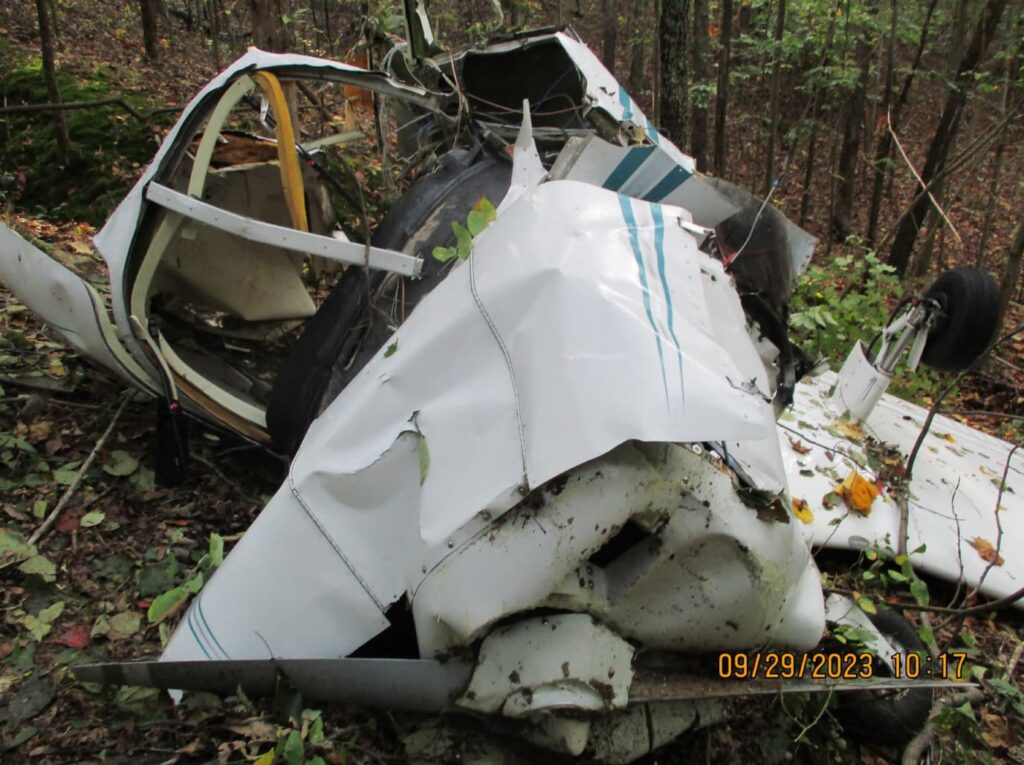 Heavily damaged fuselage and landing gear of the Piper PA-28 lying in forest terrain, with twisted metal and detached components scattered on the ground.