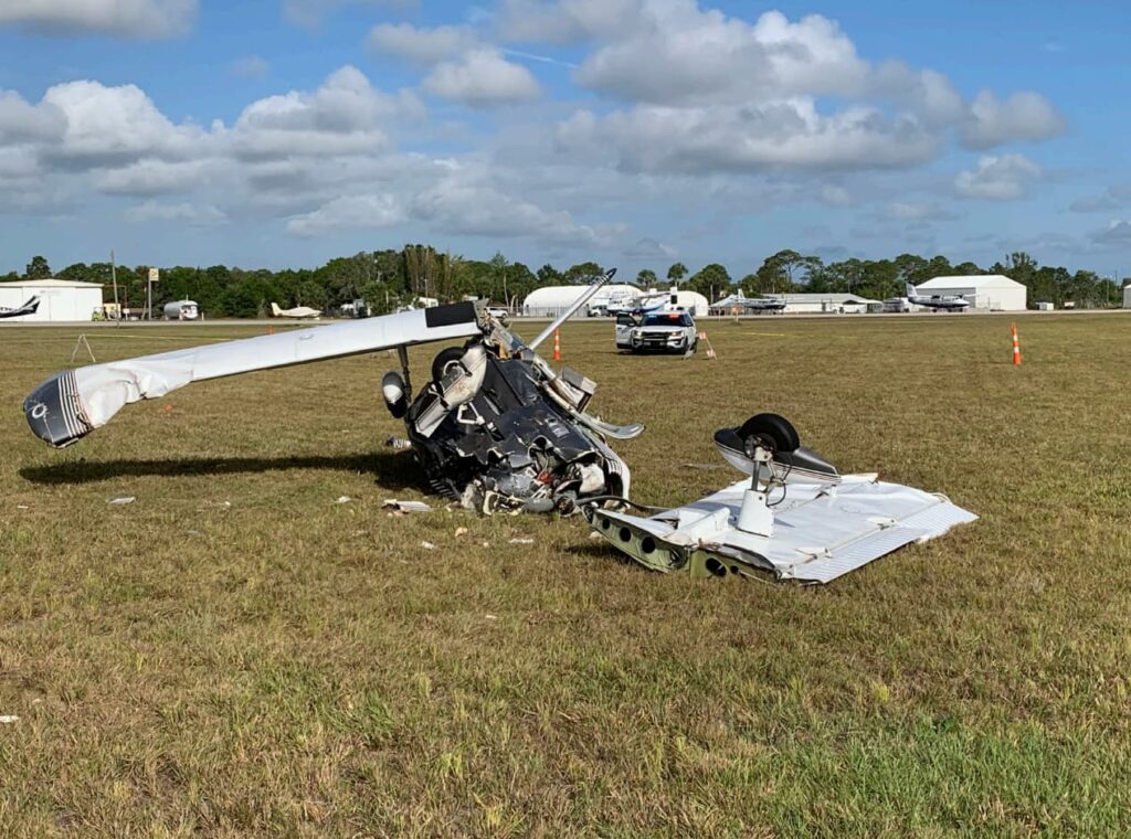 Close-up of the wrecked Piper PA-32-260 lying inverted in grass. The nose and cockpit area are crushed, with the windshield destroyed and debris scattered around. The fuselage is buckled but the tail remains attached, with hangars and cars visible in the background.