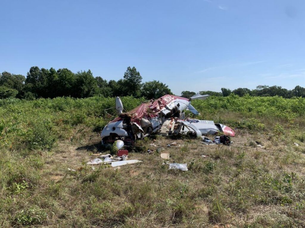 Frontal view of the same Piper PA-32 crash site in a field, showing the destroyed nose section, broken propeller, collapsed fuselage, and scattered personal items amid the grass.